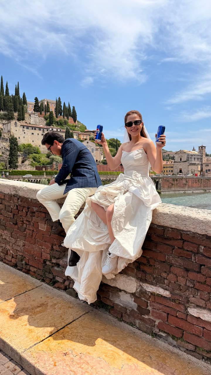 Stylish couple on a brick bridge holding energy drinks with historic cityscape behind.