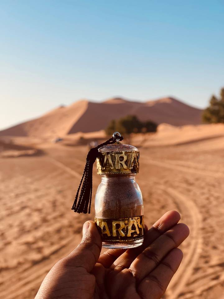 Decorative bottle filled with Sahara sand held against blurred backdrop of sun-lit dunes.