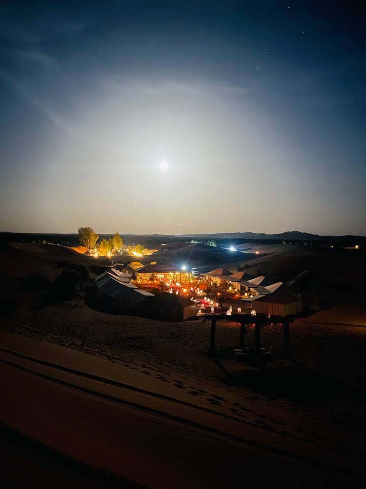 Illuminated luxury desert camp nestled between moonlit dunes under starry sky.