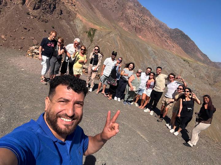 Large tour group posing on a windy mountain pass with guide taking selfie in foreground.