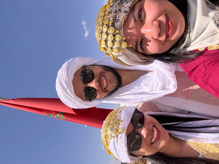 Three travelers in traditional headscarves smiling beneath Moroccan flag against bright blue sky.