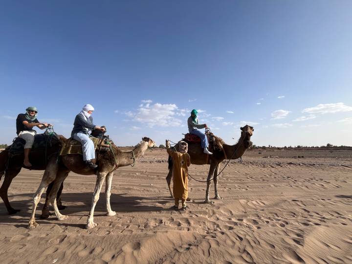 Travelers mounted on camels led by a guide in traditional attire across a sandy desert plain