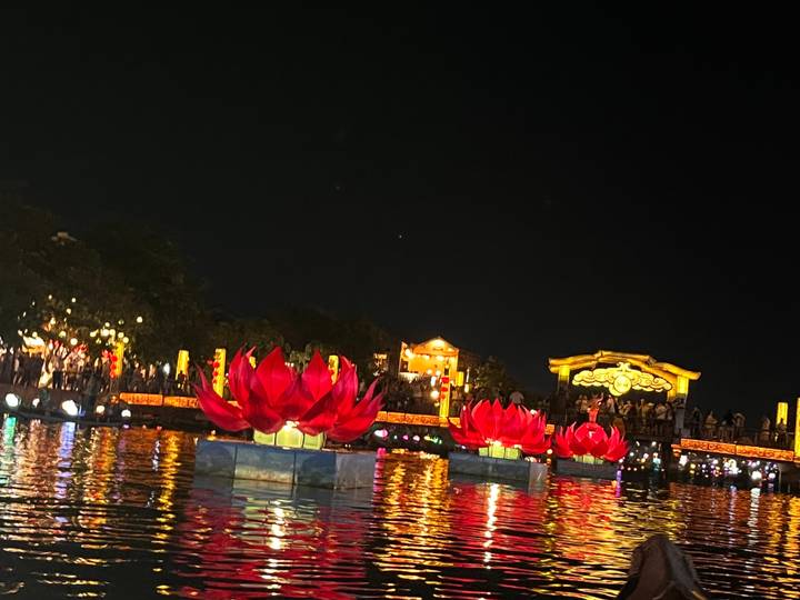 Illuminated red lotus lanterns floating on a river at night with crowds on a brightly lit bridge in the background.