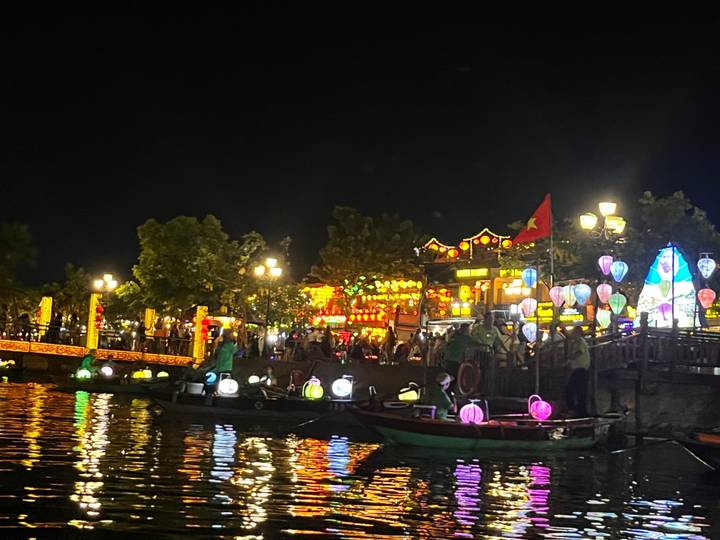 Riverfront scene at night with colorful lantern-lit boats and glowing buildings reflecting on the water.