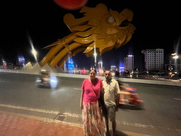 Couple standing at night on bridge beneath the lit golden dragon installation with blurred traffic passing by.