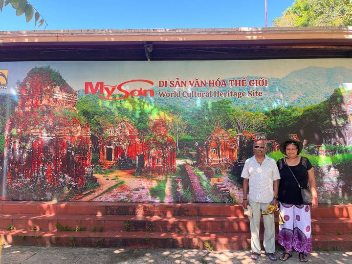 Tourists standing beside a colorful billboard for My Son World Cultural Heritage Site with forested hills depicted behind.