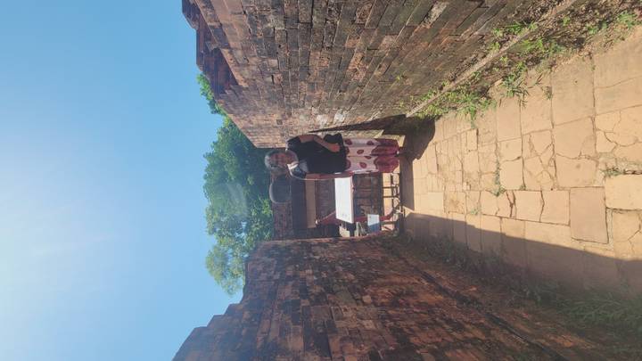 Woman standing between ancient brick walls on a sunlit stone path at an archaeological site.