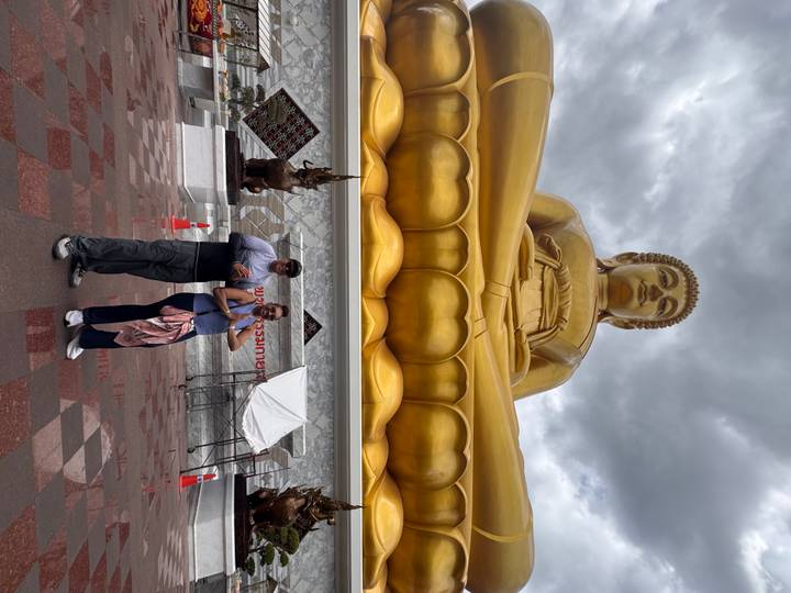 Travellers stand in prayer pose beneath a towering golden seated Buddha statue under dramatic grey clouds.