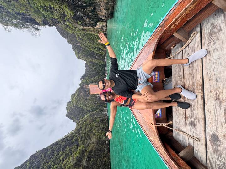 Smiling couple sitting on the bow of a long-tail boat in turquoise water surrounded by limestone cliffs.