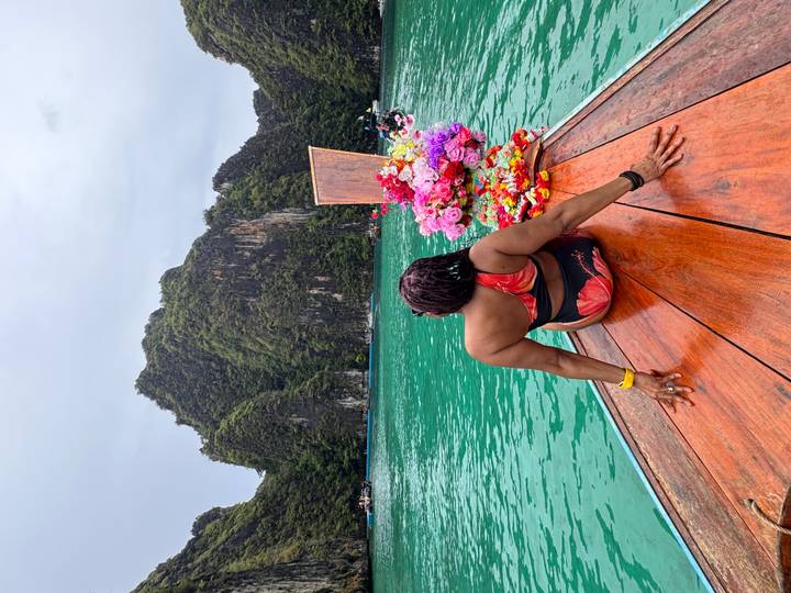 Woman relaxing on the wooden prow of a decorated long-tail boat facing emerald waters and lush cliffs.