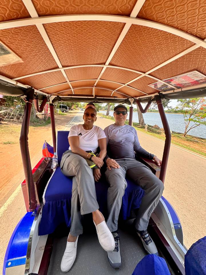 Couple riding in a blue cushioned tuk-tuk along a rural road lined with trees and water.