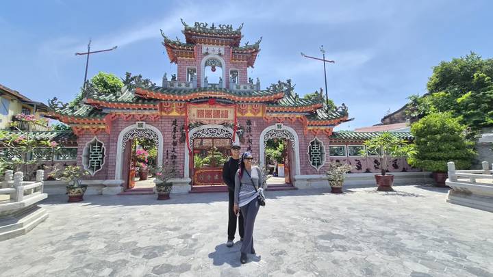 Couple standing in front of an ornate pink Chinese assembly hall gate under clear blue skies in Hoi An.