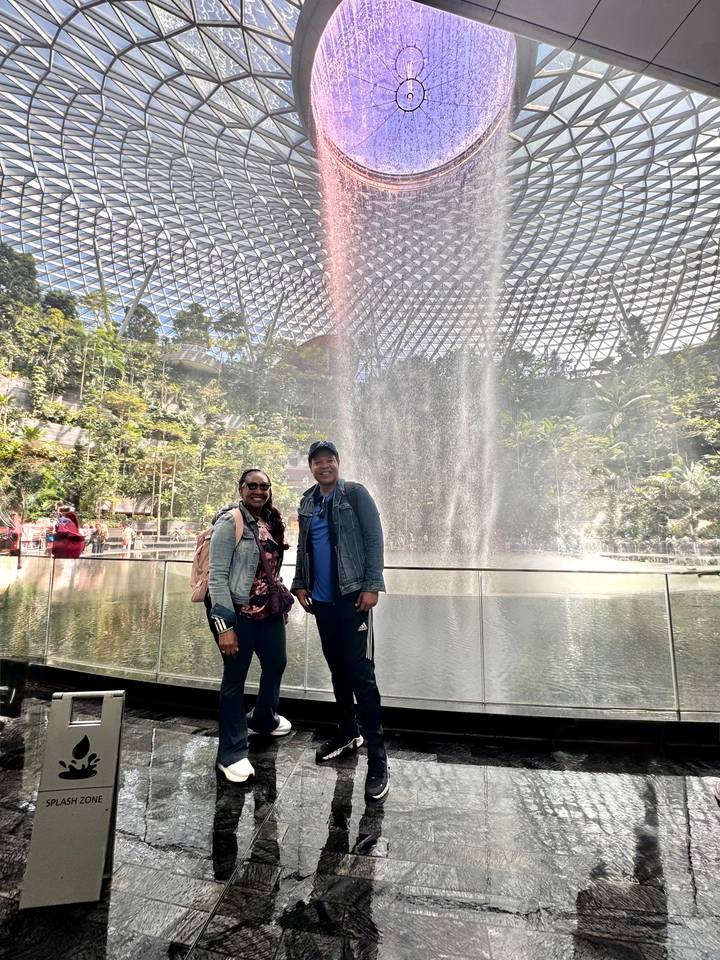 Travellers stand in front of the misty indoor waterfall under the glass dome of Jewel Changi Airport.