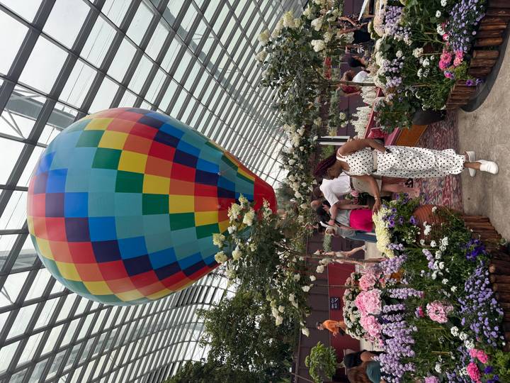 Indoor flower conservatory scene featuring a colorful hot-air balloon installation and visitors among blooming plants.