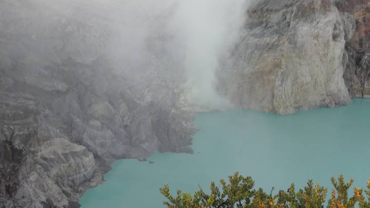 Turquoise acidic crater lake emits thick white sulphur steam from rugged volcanic walls