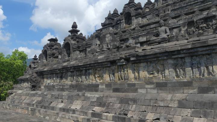 Side view of Borobudur’s stone terraces with carved reliefs and Buddha statues under clouds