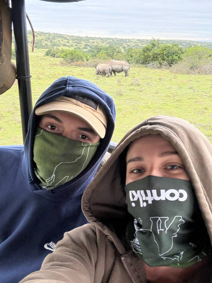 Two masked travellers take a close-up selfie while bundled in hoodies inside an open vehicle.