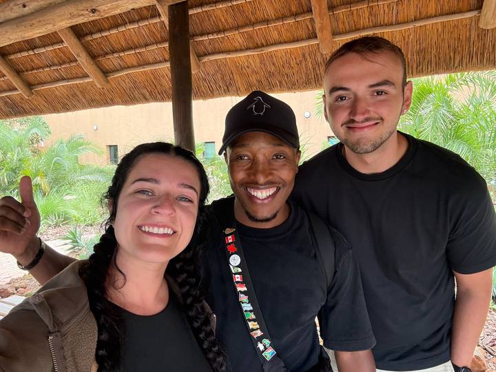 Three smiling travellers stand under a thatched roof with tropical plants behind them.