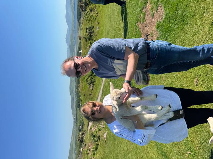 Smiling couple gently hold and feed a lamb on a sunny green hillside farm in Ireland.