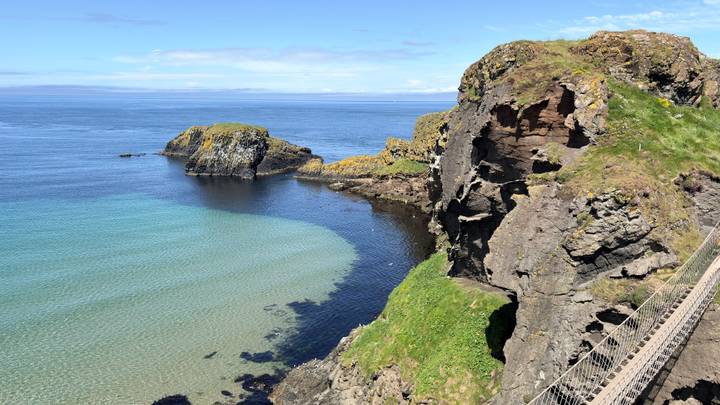 Clear turquoise cove beside rugged cliffs and a rope bridge viewpoint on a sunny day