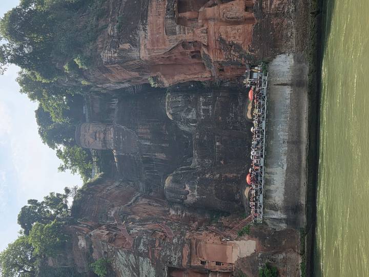Massive seated stone Buddha carved into a red cliff towering above a river with tourists on a viewing platform.