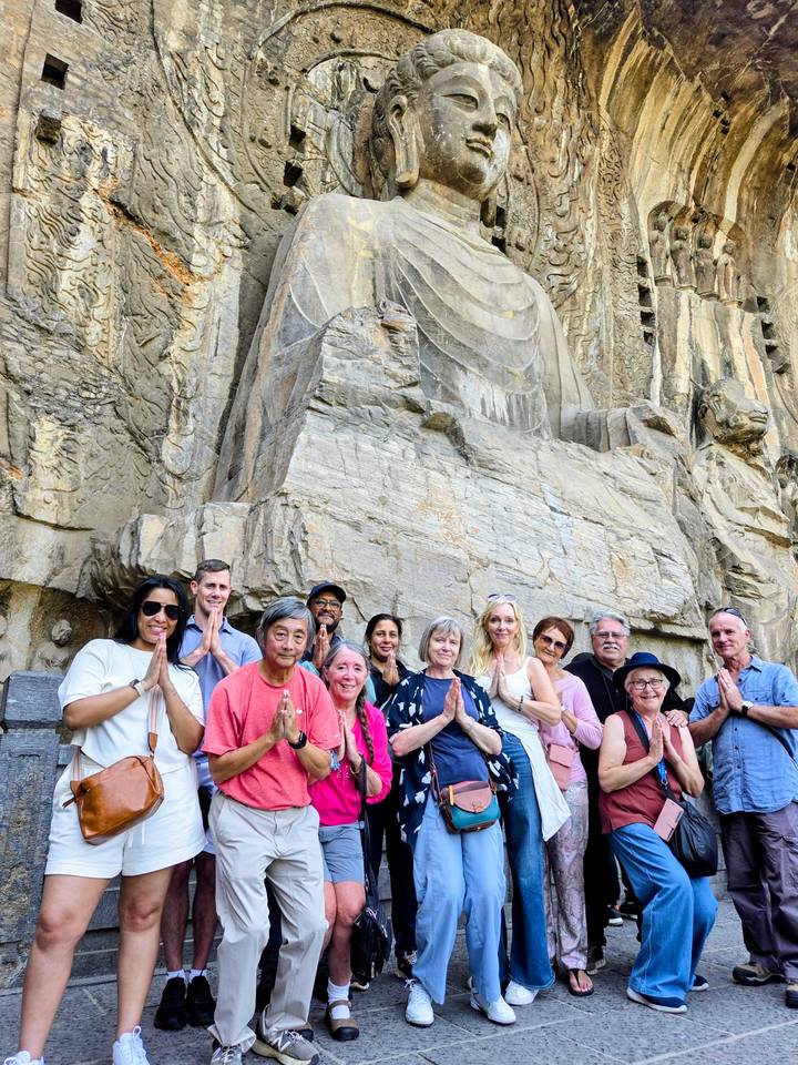 Tour group poses with prayer hands in front of a large ancient stone carving of a Buddha figure.