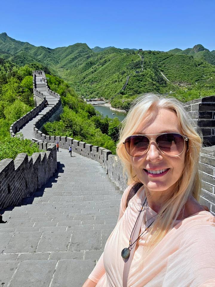 Woman takes a selfie on the battlements of the Great Wall overlooking green hills and a river bend.