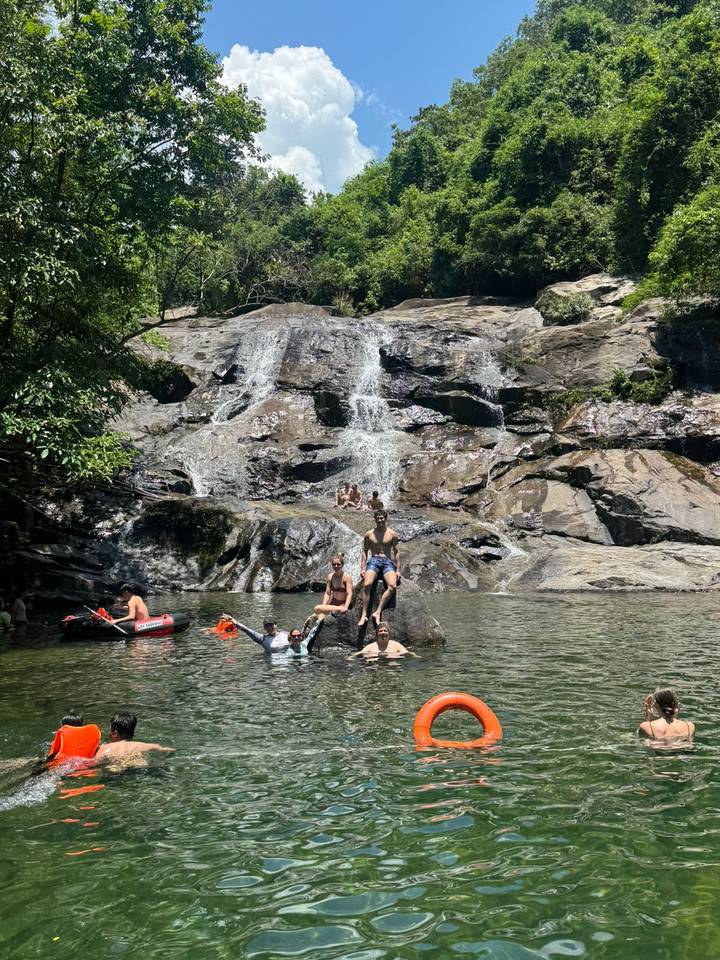 Travelers swimming and posing on rocks at a jungle waterfall pool with kayaks nearby.