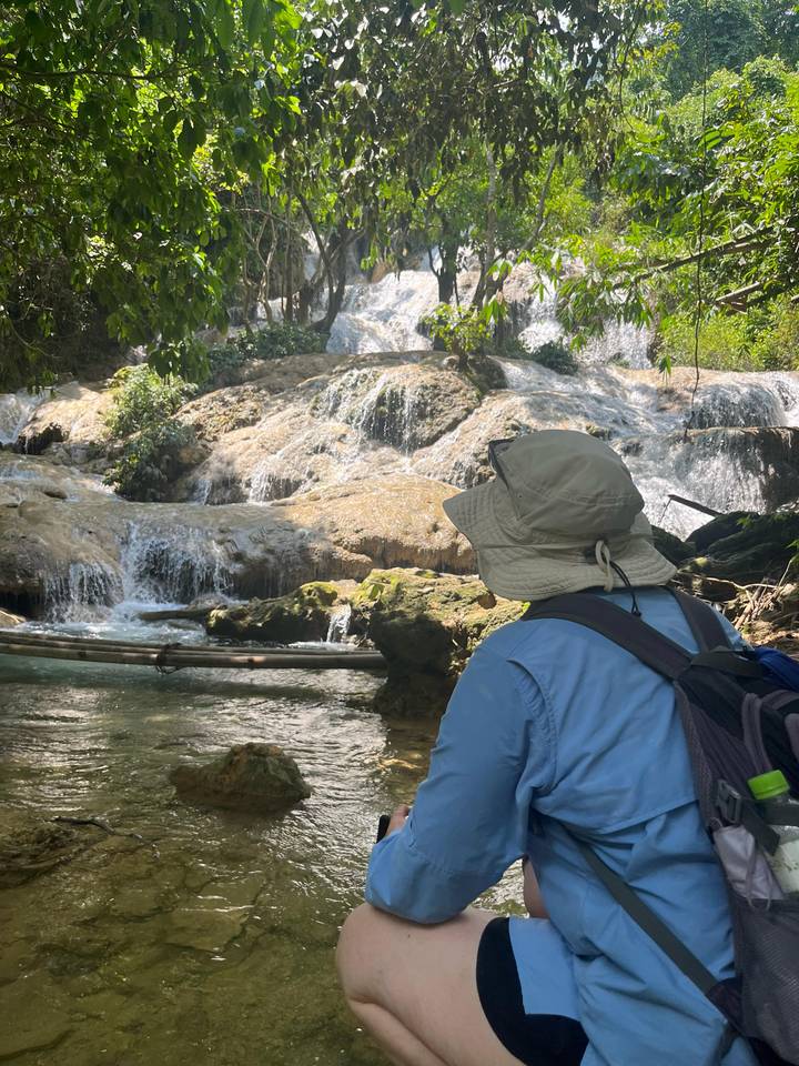 Traveler wearing a sunhat observing a tiered jungle waterfall from a shaded viewpoint.