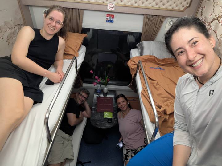 Four travelers smiling inside an overnight train cabin with bunk beds and a small table.