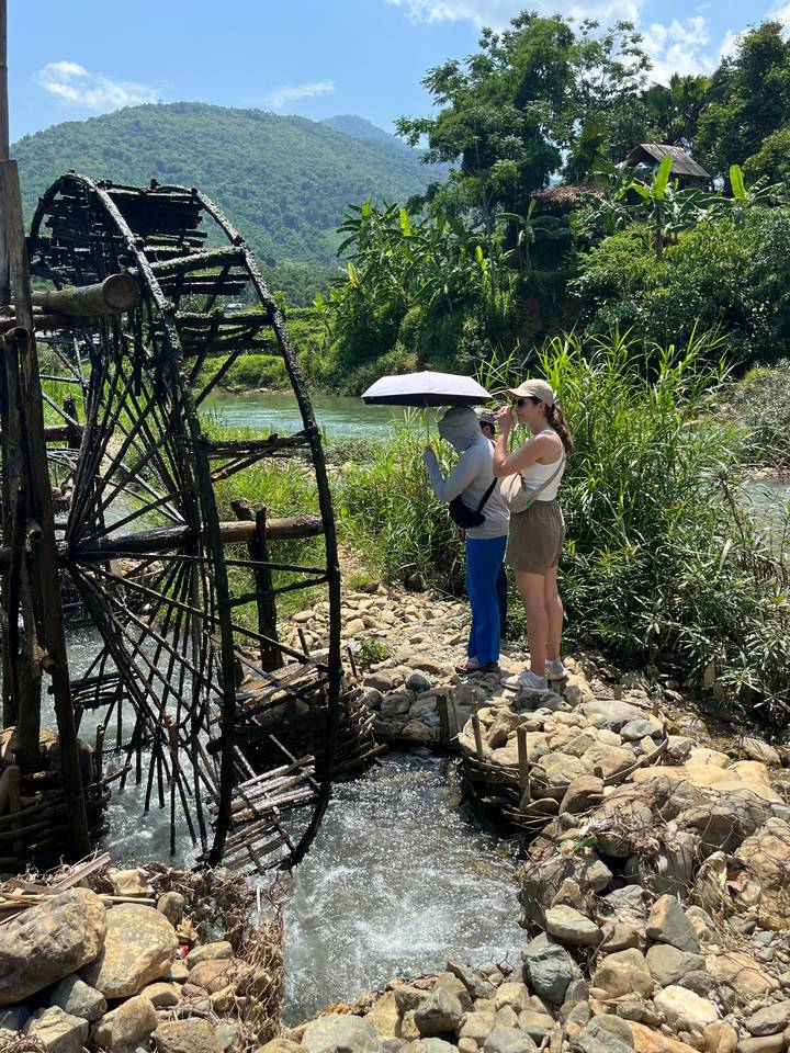 Two travelers observing a traditional wooden water wheel beside a green river while shading with an umbrella.
