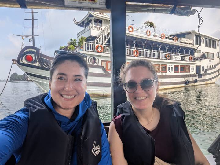 Two women wearing life vests smiling on a boat with a large white cruise ship behind in Halong Bay.