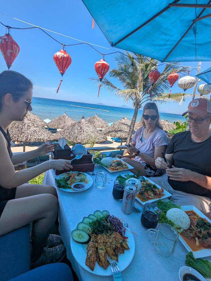 Family enjoying Vietnamese dishes at a beachfront restaurant under palm trees and thatched umbrellas.