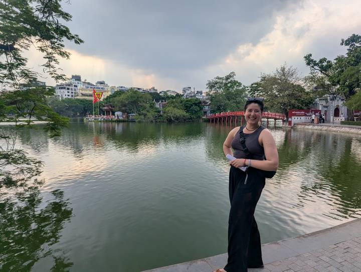 Smiling traveler standing by Hoan Kiem Lake with red bridge and city skyline in background.