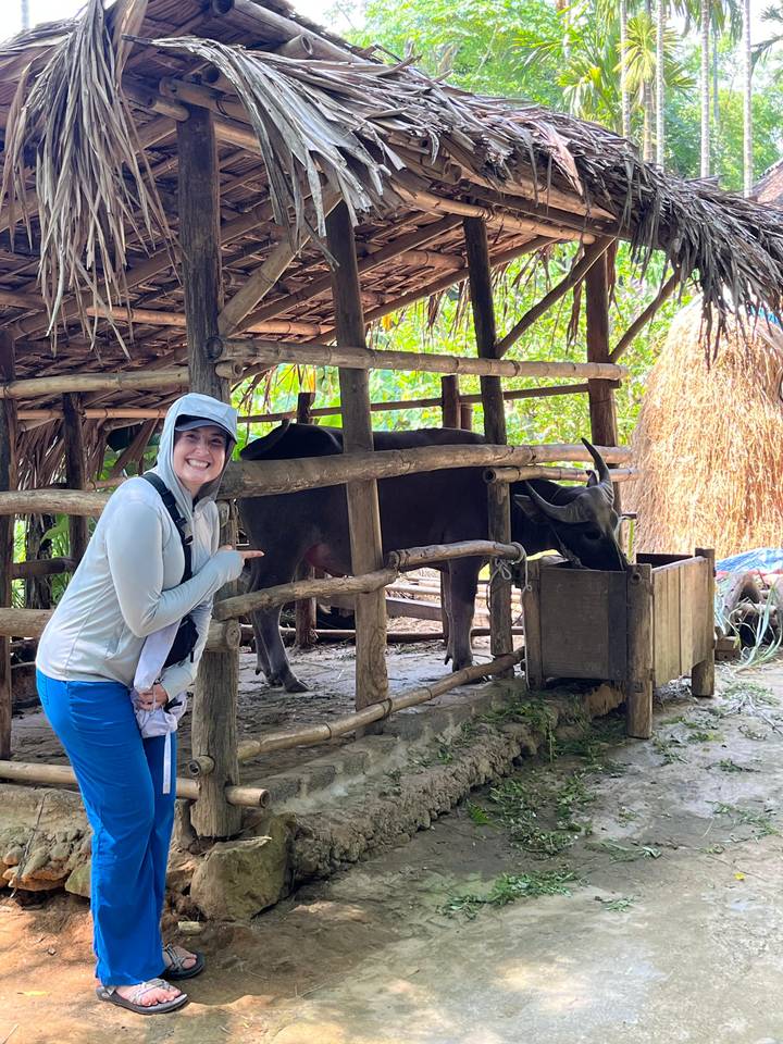 Traveler in sun hat smiling and pointing at a water buffalo feeding inside a rustic shelter.