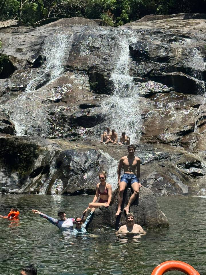 Travelers cooling off and posing on rocks and in water at the base of a cascading waterfall.