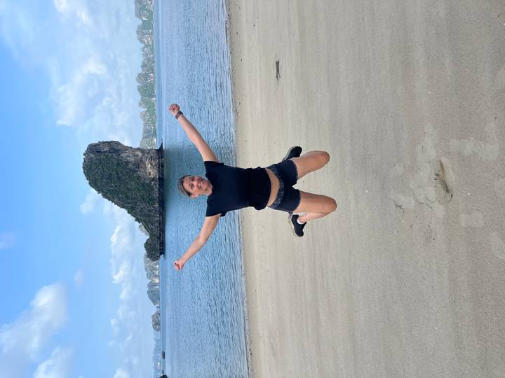 Smiling woman jumping mid-air on a quiet sandy beach with limestone island stacks in the background.