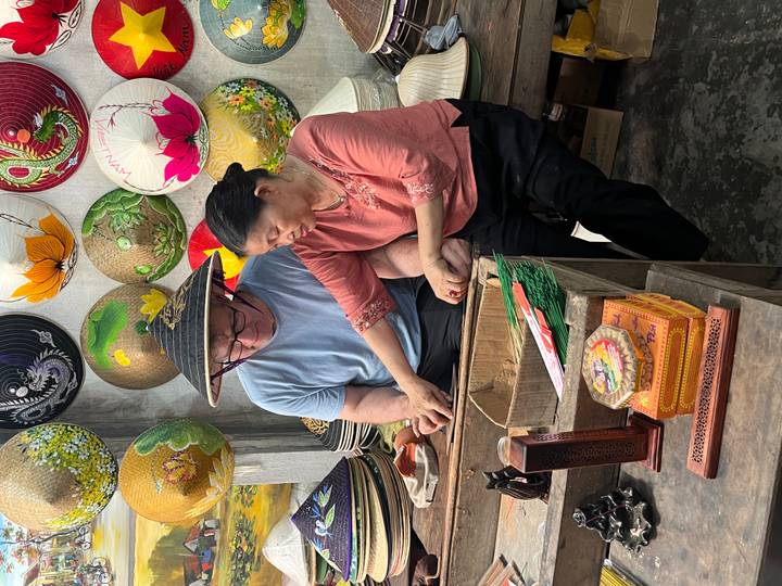 Local artisan guiding a tourist in crafting a traditional Vietnamese conical hat among colorful finished hats.
