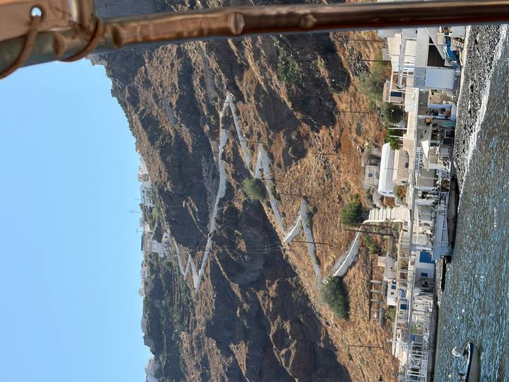 Steep zig-zagging path climbs a rocky cliffside above whitewashed buildings on the Aegean coast.