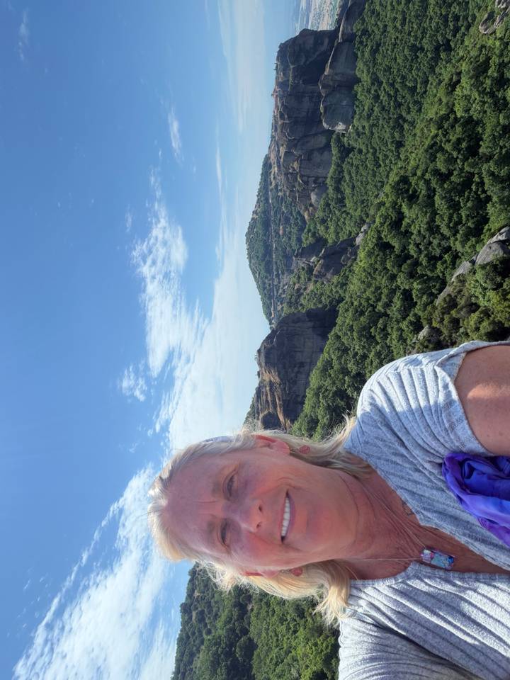 Partial selfie with expansive green valley and rocky Meteora peaks in background.