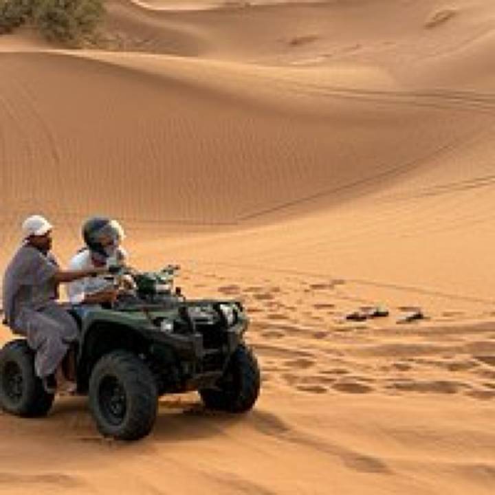 Grainy photo of two riders on a quad bike speeding over sand dunes.