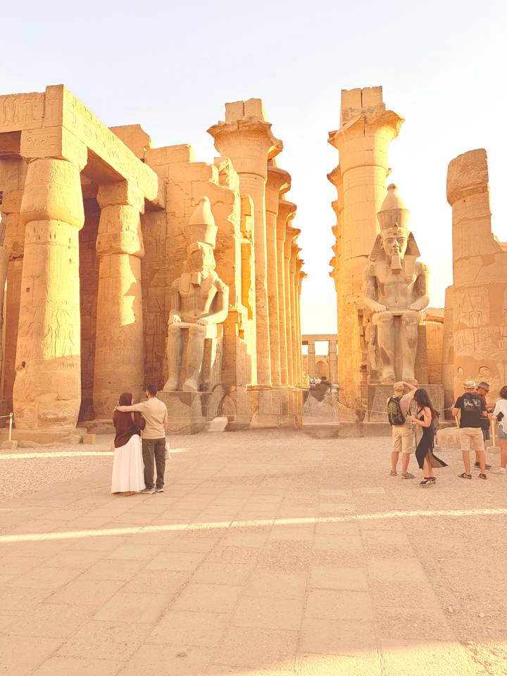Visitors wander between colossal statues and sunlit columns of Luxor Temple at golden hour.
