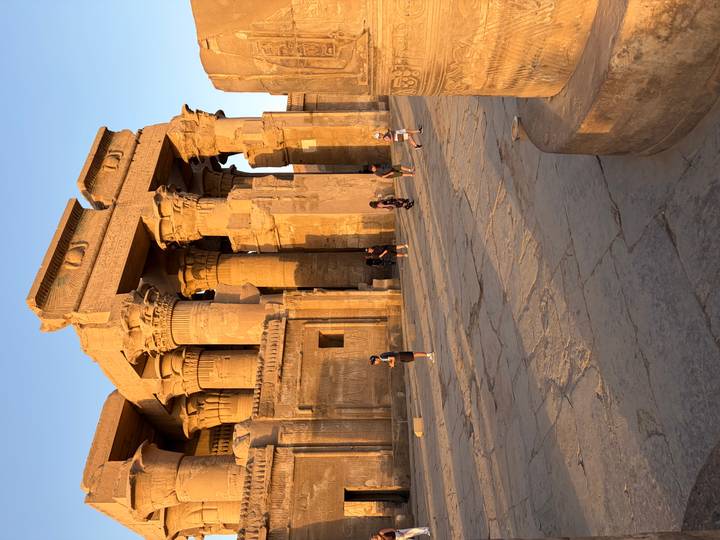 Tourists explore the impressive sandstone columns and carvings of an ancient Egyptian temple in warm afternoon light.