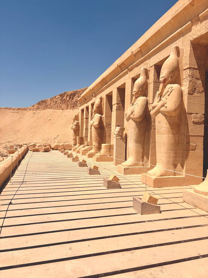 Row of monumental sandstone statues guarding a terrace against a backdrop of desert cliffs and blue sky.