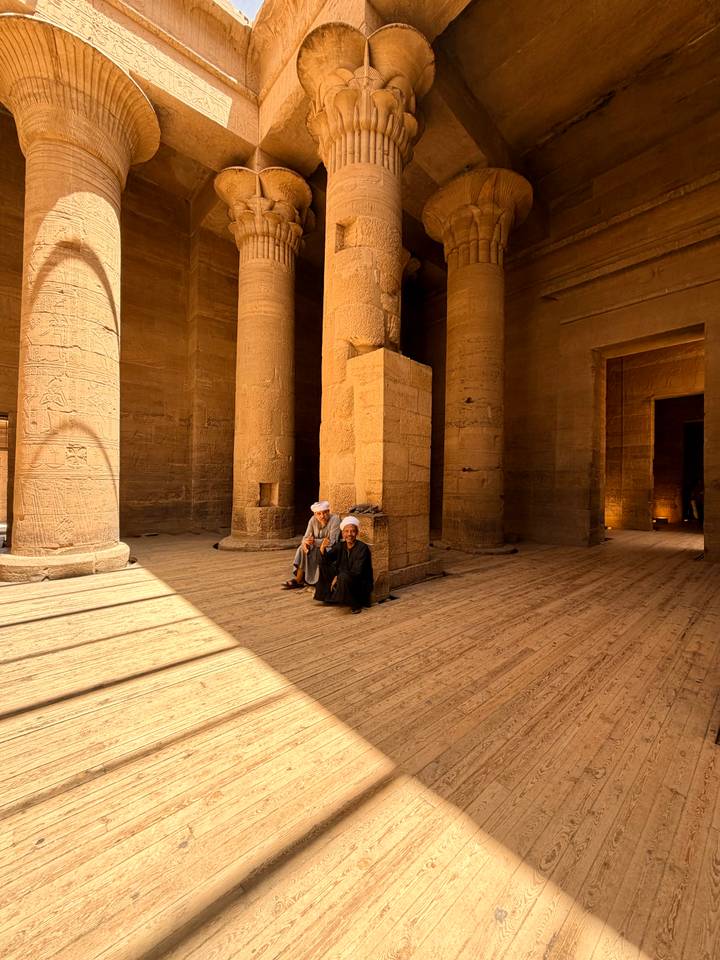 Two men in traditional robes rest inside a dimly lit hall of an ancient Egyptian temple with carved columns.