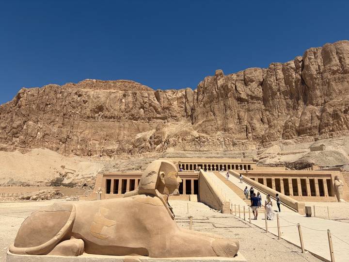 A sphinx statue overlooks tourists ascending the grand staircase of Hatshepsut Temple set beneath towering cliffs.