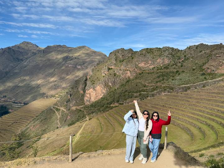 Three women celebrate on a hilltop above sweeping Inca agricultural terraces and green mountains under a bright blue sky.