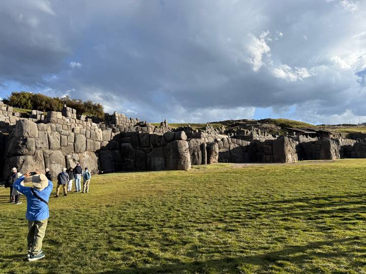 Wide lawn fronts the massive stone walls of the Sacsayhuaman fortress as tourists explore under dramatic clouds.