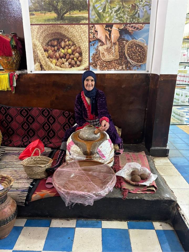 Woman demonstrates traditional argan grinding while smiling inside a cooperative stall.