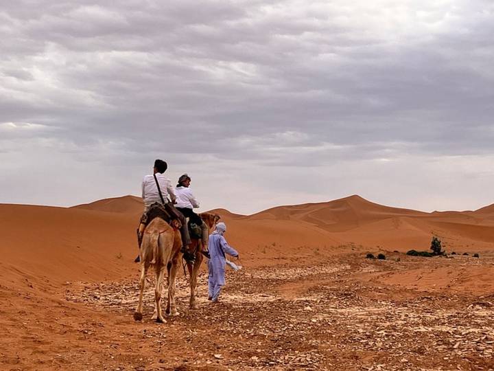 Pair of travellers ride a camel led by a guide across rust-coloured dunes under an overcast sky.